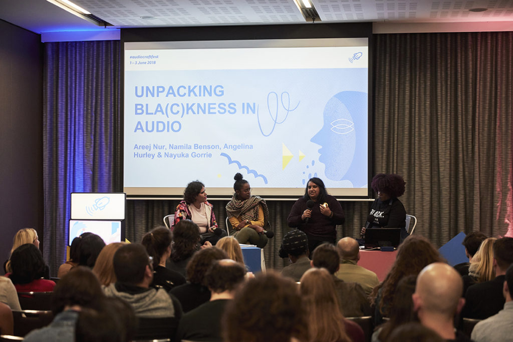 Photograph of four women speaking under a slide that reads 'Unpacking Bla(c)kness in Audio' and 'Areej Nur, Namila Benson, Angelina Hurley and Nayuka Gorrie'. Four full rows of seated attendees are listening.