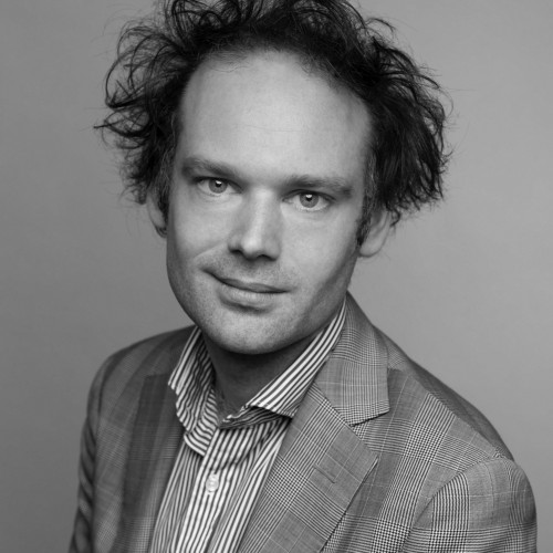 Black and white photograph of Erik Jensen, a caucasian man looking at the camera with a slight smile, tousled hair, an open-collared striped shirt and a suit jacket